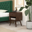 Wooden nightstand next to a green upholstered bed with a plant and decorative items.