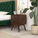 Wooden nightstand with a lamp and mug next to a green upholstered bed in a bedroom setting.
