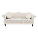 Front view of a Skein traditional three-seater sofa in light cream velvet, featuring a button-tufted camelback, two round throw pillows, rolled arms with nailhead trim, and dark turned legs in a white background.