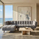 Adele 103-inch ash grey fabric left-facing sectional sofa featuring grid-tufted upholstery and tapered wood legs, styled in a sunlit modern living room with large ocean-view windows and a low travertine coffee table.