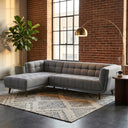 Adele 103-inch ash grey fabric left-facing sectional sofa with grid-tufted upholstery and tapered wood legs, styled on a patterned area rug in an industrial loft living room featuring exposed brick walls, a brass arc floor lamp, and a potted fiddle leaf fig plant.