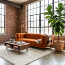 Modern mid-century Adele 85-inch burnt orange velvet sofa with tufted upholstery, styled in an industrial loft living room featuring exposed brick walls, large black-paned grid windows, a walnut wood coffee table, a textured cream rug, and a tall potted fiddle-leaf fig plant.