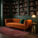Adele 85-inch channel-tufted burnt orange velvet sofa with two emerald green pillows, placed on a red patterned Persian rug in a moody library with dark green walls, floor-to-ceiling bookshelves, and a brass side table with a glowing dome lamp.