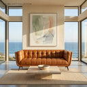 Adele 85-inch cognac top-grain leather sofa with grid-tufted upholstery, styled in a modern, sunlit living room with floor-to-ceiling windows offering a panoramic ocean view, featuring a travertine coffee table and abstract artwork.