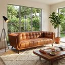 Adele 85-inch cognac top-grain leather sofa with tufted upholstery and tapered wood legs in a bright, modern living room featuring a large window, potted plant, coffee table, and tripod floor lamp.