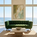 Modern Adele 85" green velvet tufted sofa with wood legs, set in a bright minimalist living room with a travertine coffee table, jute rug, abstract green and gold wall art, and floor-to-ceiling windows showcasing a scenic ocean view.