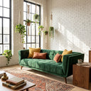 Modern green velvet channel-tufted sofa with mustard yellow and rust orange accent pillows in a bright living room featuring a white brick wall, large windows, hanging macrame plants, and a patterned area rug.