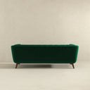 Rear view of the Adele 85-inch emerald green velvet sofa with button tufting and mid-century tapered wooden legs, isolated on a beige background.