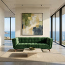 A modern coastal living room featuring the Adele 85 green velvet tufted sofa on a beige area rug, a travertine coffee table, and a large abstract painting, flanked by floor-to-ceiling windows with a scenic ocean view.