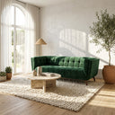 Emerald green velvet tufted sofa (Adele 85-inch) in a bright, modern living room with a round travertine coffee table, textured cream area rug, large arched window with sheer curtains, and an olive tree in a terracotta pot.