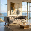 Adele 85-inch tufted blue velvet sofa in a modern living room with floor-to-ceiling windows overlooking the ocean, featuring an abstract painting on the wall and a low marble coffee table on a jute rug.