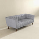 Three-quarters view of the Adele 85-inch blue velvet sofa with button tufting on the back, channel-tufted seat cushions, and tapered dark wood legs, isolated on a beige background.