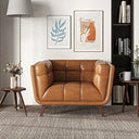 Adele cognac brown leather accent armchair featuring deep grid-tufted cushions, wide rounded armrests, and tapered wooden legs. It is centered in a modern living room on a neutral textured rug, flanked by a tall floor lamp and small side tables, with framed wall art and a tall bookshelf in the background.