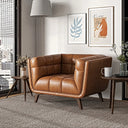 Mid-century modern cognac brown leather accent armchair with tufted, channel-stitched interior cushioning and tapered dark wooden legs, styled on a textured rug in a bright room with wooden side tables, framed art, and a window overlooking a city.