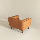 Rear three-quarter view of the Adele accent chair upholstered in cognac brown leather, highlighting its smooth curved back, vertical seam stitching, tufted inner cushioning, and tapered dark wood legs against a neutral textured background.
