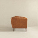 Side profile view of the Adele accent chair upholstered in warm cognac brown leather, featuring tailored panel stitching and dark wood legs against a minimalist neutral textured backdrop.