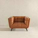 Front view of the Adele cognac leather accent chair featuring a mid-century modern design with a button-tufted backrest, plush channeled armrests, and a deep cushioned seat, supported by dark tapered wooden legs against a neutral textured backdrop.