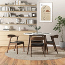 Round white-top dining table with wooden legs, surrounded by dark wood chairs with black cushioned seats on a circular woven rug in a modern dining room.
