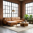 Adriana 111-inch tan genuine leather left-facing sectional sofa with tufted cushions styled in an industrial loft living room featuring exposed red brick walls, large grid windows, a brass arc floor lamp, round wooden coffee table, and potted plants.