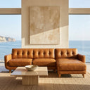 Adriana 111-inch tan genuine leather right-facing sectional sofa with button-tufted backrest and tapered wooden legs in a modern living room with ocean views.