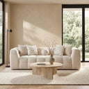 Agnes 90-inch tufted ivory chenille sofa with textured accent pillows, positioned in a modern minimalist living room in front of a warm beige plaster wall and next to large black-framed windows. A round travertine coffee table with a decorative vase and book sits on a textured cream rug in the foreground.