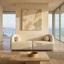 Alexis 84-inch ivory bouclé sofa with curved arms and two spherical pillows in a sunlit, modern living room with scenic coastal ocean views, a large textured neutral abstract painting, and a travertine coffee table on a woven area rug.