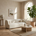 Alexis 84-inch curved ivory boucle sofa with round matching throw pillows in a minimalist living room featuring a travertine coffee table, a jute rug, and a potted fiddle leaf fig plant by an arched window.