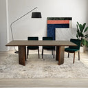 Almaza walnut dining table with angled pedestal legs, paired with dark green velvet chairs on a neutral textured rug, featuring a black arc floor lamp and modern artwork in the background.
