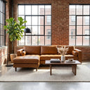 Anita 102-inch tan genuine top-grain leather left-facing sectional sofa with tufted seating and bolster pillows, set in a loft living room featuring exposed brick walls, large industrial windows, a wooden coffee table, and a potted fiddle-leaf fig.