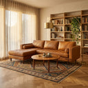 Anita 102-inch tan genuine top grain leather left-facing sectional sofa with tufted cushions and cylindrical bolster pillows set in a sunlit living room with a round wooden coffee table and patterned rug.