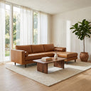 Anita 102 tan genuine top grain leather right-facing sectional sofa with tufted seat cushions and cylindrical bolster pillows in a sunlit modern living room with floor-to-ceiling windows, sheer curtains, a wooden coffee table, and a potted plant.