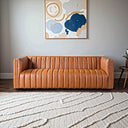 April 86-inch cognac genuine leather sofa with vertical channel tufting in a modern living room setting with abstract wall art and a textured cream area rug.