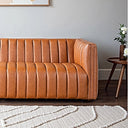 April 86-inch cognac genuine leather sofa with vertical channel tufting displayed in a modern living room on dark wood flooring next to a textured cream rug.