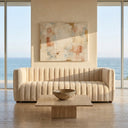 April 86-inch Mid-Century Modern channel tufted beige boucle sofa in a minimalist living room with a travertine coffee table, large abstract painting, and floor-to-ceiling windows showing a scenic ocean view.