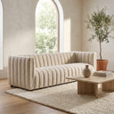 April 86-inch mid-century modern tufted beige boucle sofa with vertical channel tufting, styled on a textured cream rug in a bright minimalist living room with a travertine coffee table and a potted olive tree near an arched window.