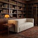 April 86 Mid-Century Modern channel-tufted beige boucle sofa in a warm library setting with walnut bookshelves, a brass library ladder, an end table with a glowing amber glass lamp, and a patterned rug.