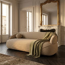 Ava 84-inch modern curved sofa in beige bouclé fabric with a matching round pillow and draped olive green throw blanket, situated in a Parisian-style living room featuring a white marble fireplace, ornate gold gilded mirror, and herringbone hardwood floors.