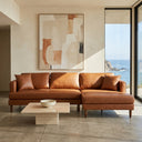Bayley 107-inch cognac brown vegan eco-leather right-facing sectional sofa with chaise, wooden legs, and matching pillows in a modern sunlit living room with an ocean view.
