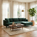 Bayley 107-inch dark green velvet left-facing sectional sofa with matching accent pillows set in a bright, modern living room with a wooden coffee table, plush shag rug, brass arc floor lamp, and potted fiddle-leaf fig plant.