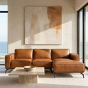 Berry 102-inch cognac brown vegan eco-leather right-facing sectional sofa with black metal legs in a modern sunlit living room featuring ocean views, a low travertine coffee table, and large abstract wall art.