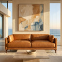 Brynn 93-inch mid-century modern tan genuine top-grain leather sofa in a luxury coastal living room with floor-to-ceiling windows overlooking the ocean. An abstract art painting in beige, brown, and blue hangs on the wall behind the sofa, and a travertine coffee table is in the foreground.