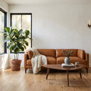 Brynn 93-inch mid-century modern tan genuine top-grain leather sofa draped with a cream knit throw blanket, styled in a bright living room with a triangular wood coffee table and a large fiddle-leaf fig plant by a window.