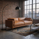 Brynn 93-inch Mid-Century Modern tan genuine top-grain leather sofa in an industrial loft apartment with a red brick wall, a black arched floor lamp, a patterned area rug, and large multi-pane windows showing a city skyline.
