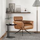 Modern tan genuine leather swivel armchair with black metal legs, styled in a bright living space on a textured grey rug. Beside the chair is a round black side table with a coffee cup and green foliage in a vase. In the background, a floating wooden shelf displays framed art and decor, complemented by a tall floor lamp with a pleated shade.