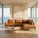 Carla 109-inch tan genuine leather right-facing sectional sofa with tapered wooden legs in a modern sunlit living room with ocean views.