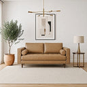 Mid-century modern tan genuine leather loveseat with bolster pillows, tufted seat cushion, and wooden legs in a bright living room alongside a potted tree, framed geometric art, a brass chandelier, and a side table with a lamp.
