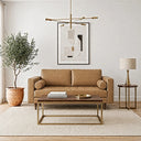 A mid-century modern tan leather loveseat with bolster pillows in a bright living room, paired with a wood-and-brass coffee table, a potted olive tree, a brass table lamp on a side table, framed geometric wall art, and a brass chandelier.
