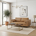 Tan genuine leather mid-century modern loveseat with bolster pillows and tufted seat in a bright living room, paired with a wood and brass coffee table, potted olive tree, framed geometric wall art, and a brass sputnik chandelier.

