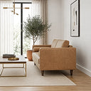 Side view of a tan genuine leather loveseat with bolster pillows and dark wood legs in a bright living room, beside a wood-and-brass coffee table, neutral area rug, potted olive tree, and framed abstract art.
