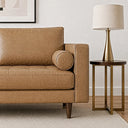 Close-up of the right side of a tan genuine leather mid-century modern loveseat with a tufted seat, bolster pillow, square track arm, and tapered wooden legs, next to a brass and wood end table topped with a brass table lamp.
